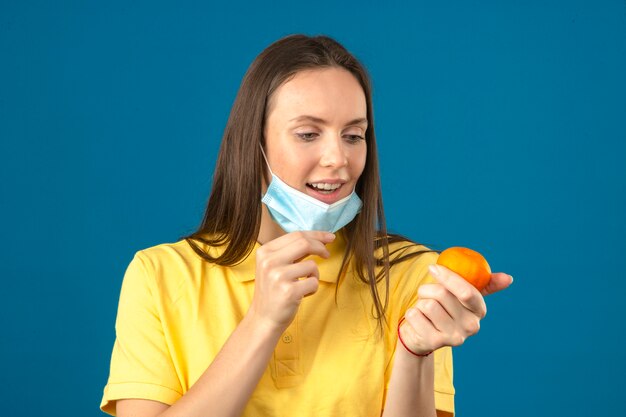 Joven vistiendo polo amarillo quitándose la máscara médica protectora y sosteniendo naranja mandarina mirando cítricos en mano sobre fondo azul.