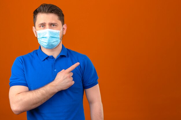 Joven vistiendo polo azul en máscara protectora médica apuntando con el dedo hacia el lado de pie sobre la pared naranja aislada