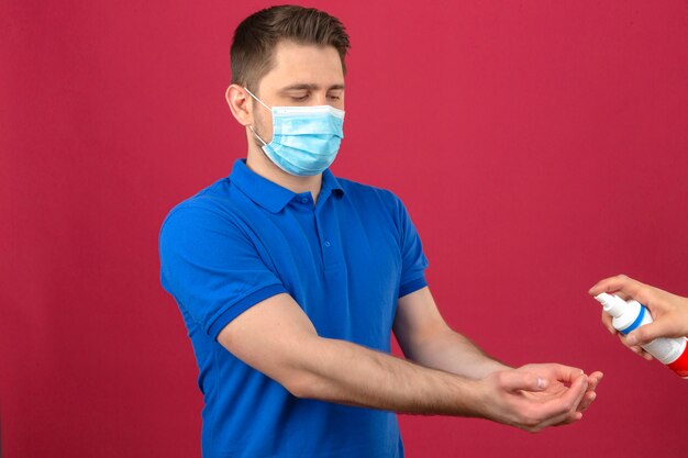 Joven vistiendo polo azul en máscara protectora médica estirando sus manos para desinfectante desinfectando sus manos sobre pared rosa aislado