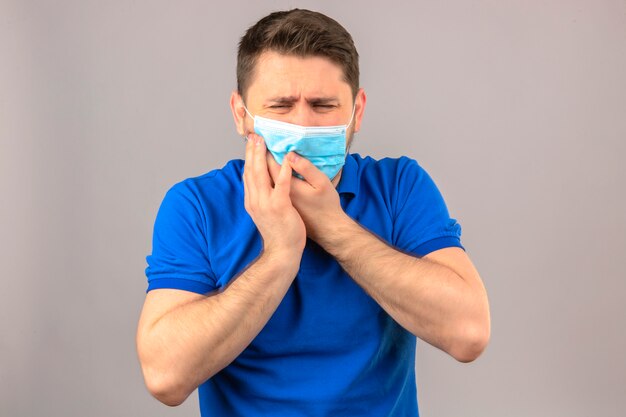 Joven vistiendo polo azul en máscara protectora médica mirando mal tocar la mejilla que sufre de dolor de muelas sobre la pared blanca aislada