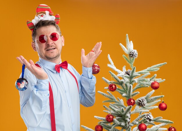 Joven vistiendo tirantes pajarita en el borde con santa y gafas rojas de pie junto al árbol de navidad mirando confundido sosteniendo juguetes para árbol sobre fondo naranja