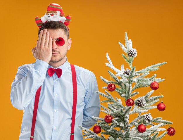 Joven vistiendo tirantes pajarita en el borde con santa y gafas rojas de pie junto al árbol de navidad que cubre un ojo con la mano mirando a la cámara sobre fondo naranja