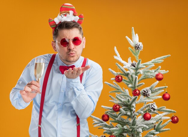 Joven vistiendo tirantes pajarita en el borde con santa y gafas rojas de pie junto al árbol de navidad sosteniendo una copa de champán feliz y alegre que sopla un beso sobre fondo naranja
