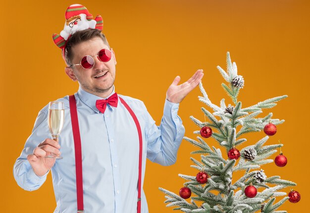 Joven vistiendo tirantes pajarita en el borde con santa y gafas rojas de pie junto al árbol de navidad sosteniendo una copa de champán feliz y alegre sobre fondo naranja