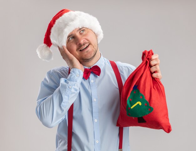 Joven vistiendo tirantes pajarita con gorro de Papá Noel con bolsa de santa claus llena de regalos mirando hacia arriba sonriendo feliz y positivo de pie sobre fondo blanco.