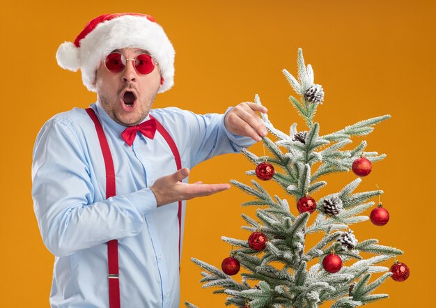 Joven vistiendo tirantes pajarita con gorro de Papá Noel y gafas rojas de pie cerca del árbol de Navidad feliz y asombrado juguete colgando en el árbol sobre la pared naranja