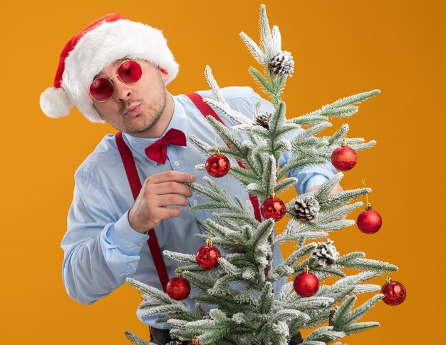 Joven vistiendo tirantes pajarita con gorro de Papá Noel y gafas rojas de pie detrás del árbol de Navidad feliz y asombrado juguete colgante en árbol sobre fondo naranja