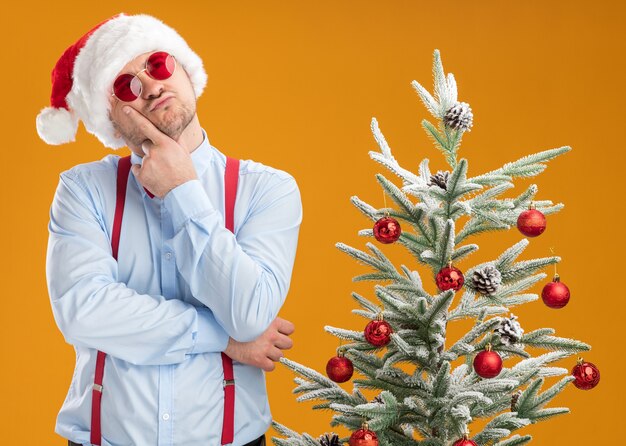 Joven vistiendo tirantes pajarita con gorro de Papá Noel y gafas rojas de pie junto al árbol de Navidad mirando hacia arriba con la mano en la barbilla pensando en la pared naranja