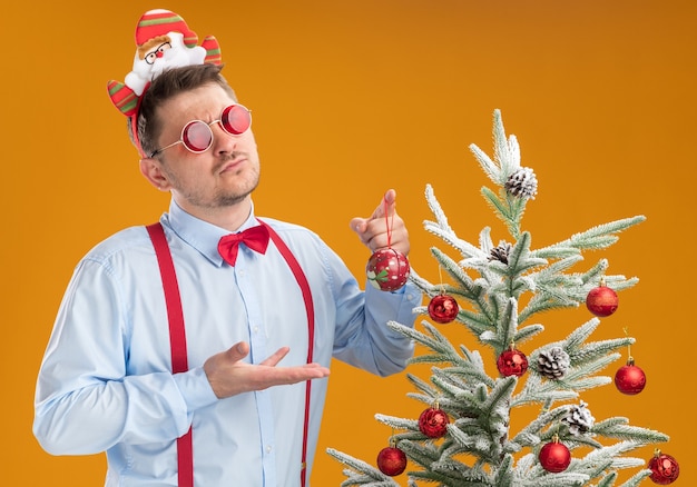 Joven vistiendo tirantes pajarita en santa sombrero y gafas rojas de pie junto al árbol de navidad sobre pared naranja