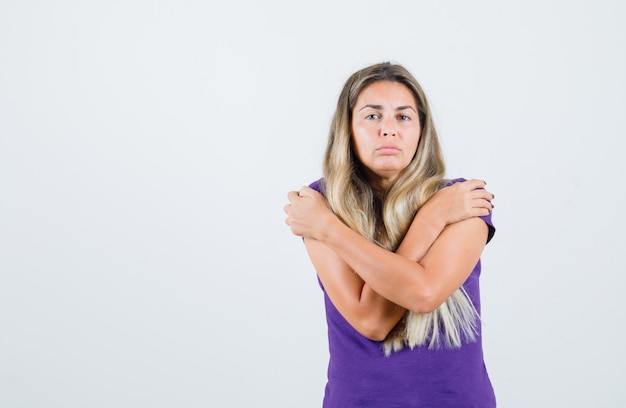 Jovencita abrazándose a sí misma en camiseta violeta y mirando triste, vista frontal.