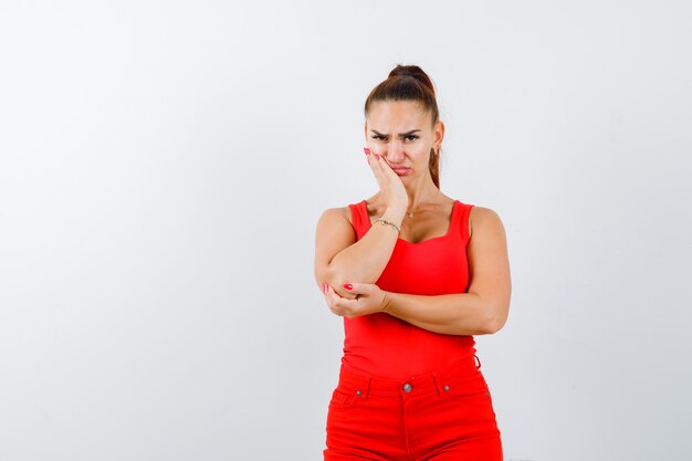 Jovencita apoyando la mejilla en la mano con camiseta roja, pantalón rojo y aspecto resentido. vista frontal.