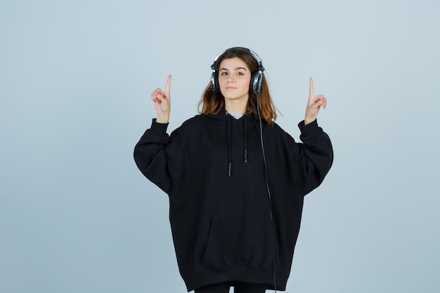 Foto gratuita jovencita apuntando hacia arriba mientras escucha música con auriculares en una sudadera con capucha de gran tamaño, pantalones y mirando confiado, vista frontal.