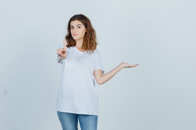 Jovencita apuntando hacia arriba mientras sostiene algo en camiseta, jeans y parece confiada. vista frontal.