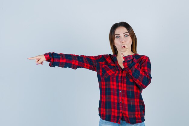 Jovencita apuntando hacia la izquierda, manteniendo la mano en la boca con la camisa a cuadros y mirando sorprendido, vista frontal.