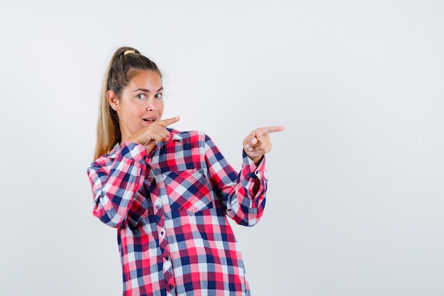 Jovencita apuntando hacia el lado derecho con camisa a cuadros y mirando vacilante, vista frontal.