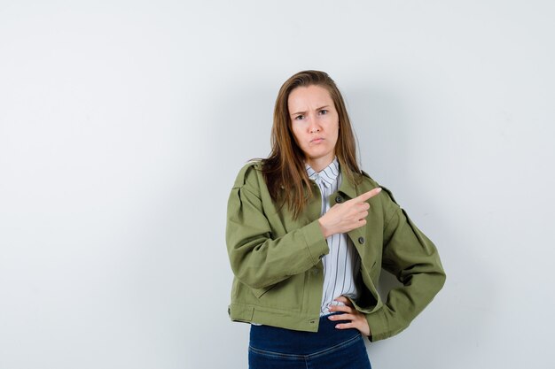 Jovencita apuntando hacia el lado derecho mientras fruncía el ceño en camisa, chaqueta y miraba vacilante. vista frontal.