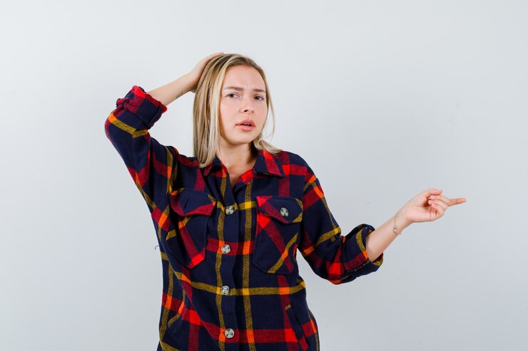 Foto gratuito jovencita apuntando hacia el lado derecho mientras sostiene la mano en la cabeza con camisa a cuadros y mira vacilante, vista frontal.