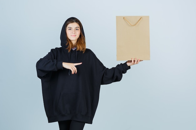 Jovencita apuntando hacia el lado derecho mientras sostiene el paquete en una sudadera con capucha de gran tamaño, pantalones y mira confiada, vista frontal.