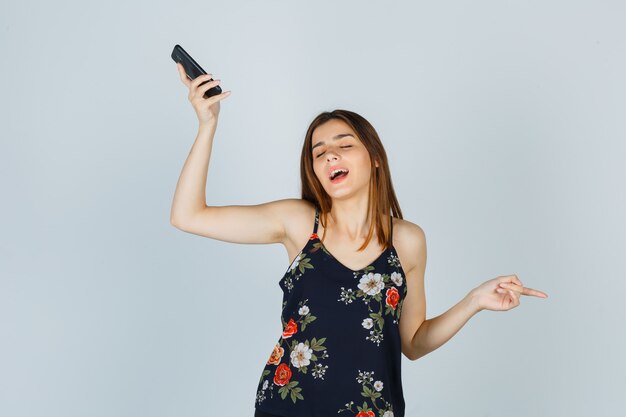 Jovencita apuntando a un lado mientras sostiene el teléfono inteligente en la blusa y mirando relajado, vista frontal.