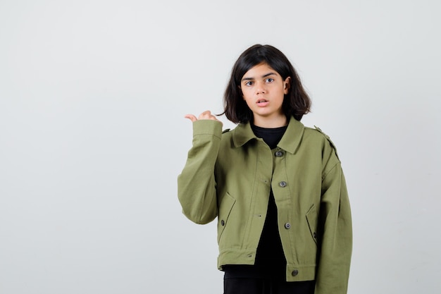 Jovencita apuntando a un lado con el pulgar en la chaqueta verde militar y mirando perplejo, vista frontal.