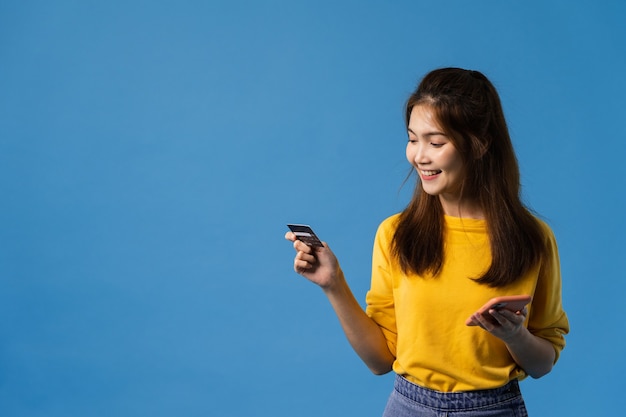 Jovencita de Asia con teléfono y tarjeta bancaria de crédito con expresión positiva, sonríe ampliamente, vestida con ropa informal y de pie aislado sobre fondo azul. Feliz adorable mujer alegre se regocija con el éxito.