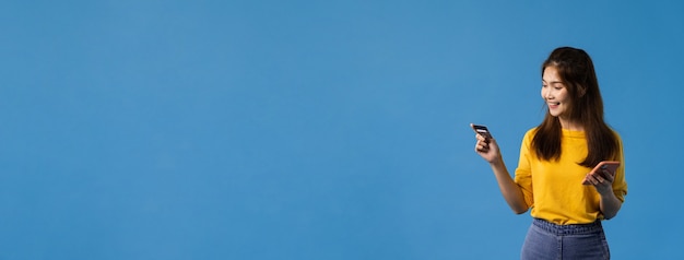 Jovencita de Asia con teléfono y tarjeta bancaria de crédito con expresión positiva, sonrisa amplia, vestida con ropa informal y aislada sobre fondo azul. Fondo de banner panorámica con espacio de copia.