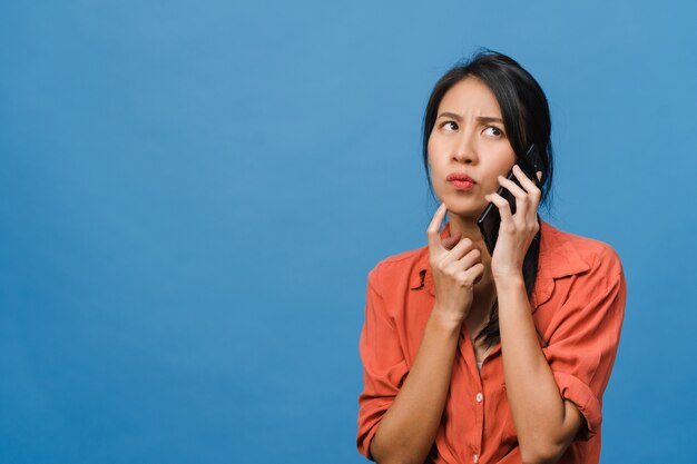 Jovencita asiática habla por teléfono con expresión negativa, gritos emocionados, llora emocionalmente enojada en ropa casual y está aislada en la pared azul con espacio de copia en blanco. Concepto de expresión facial.