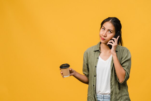 Jovencita asiática habla por teléfono y sostiene la taza de café con expresión negativa, gritos emocionados, llora enojado emocionalmente en ropa casual y se para aislado en la pared amarilla. Concepto de expresión facial.