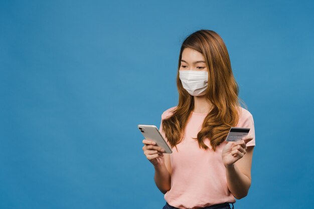 Jovencita asiática con mascarilla médica usando teléfono y tarjeta bancaria de crédito con expresión positiva, sonríe ampliamente, vestida con ropa informal y de pie aislado en una pared azul