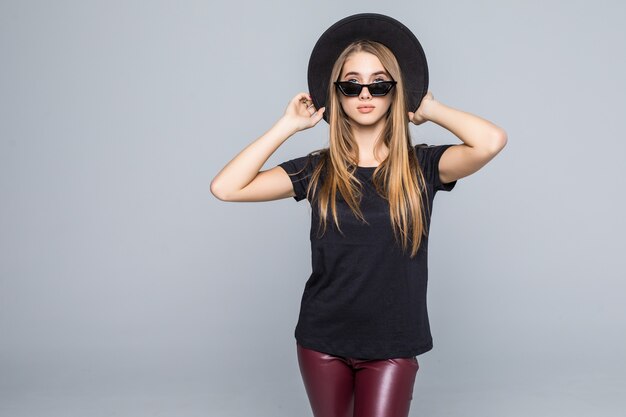 Jovencita bonita con gafas de sol brillantes vestida con sombrero negro, camiseta negra y pantalón oscuro aislado sobre fondo gris