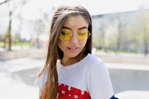 Jovencita bonita con pelo largo volando en elegantes gafas de sol cerró los ojos y sonríe al atardecer. Cerrar foto de mujer feliz en gafas de sol posando de lado al aire libre