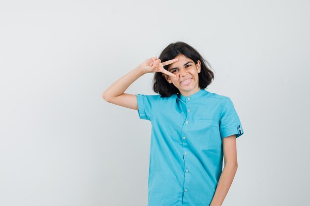 Jovencita con camisa azul mostrando el signo v sobre un ojo y luciendo alegre