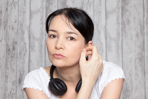 jovencita en camisa blanca escuchando música en auriculares negros en la pared gris