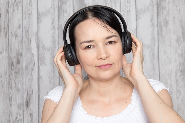 jovencita en camisa blanca escuchando música en auriculares negros en la pared gris