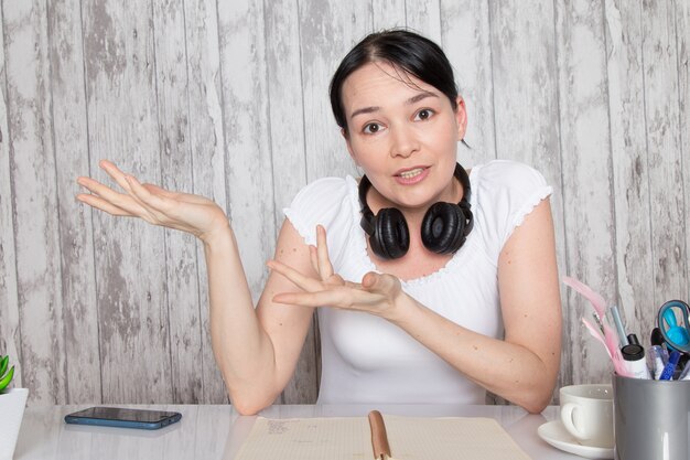 Jovencita en camisa blanca mostrando emociones con sus manos en auriculares negros sobre pared gris