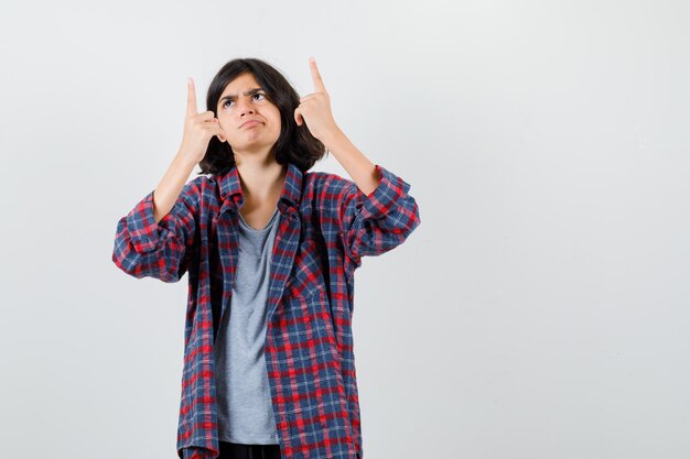 Jovencita en camisa a cuadros apuntando hacia arriba con los dedos y mirando disgustado, vista frontal.