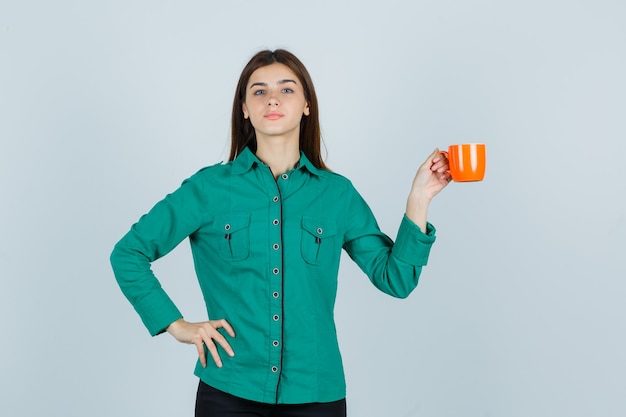 Jovencita en camisa posando mientras sostiene una taza de té naranja y mira confiada, vista frontal.