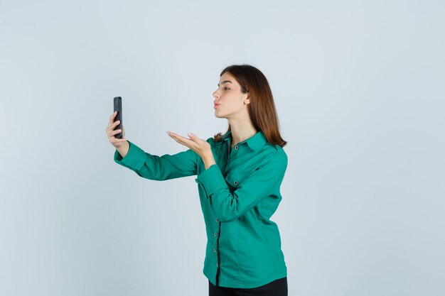 Jovencita en camisa verde enviando beso al aire mientras toma selfie en teléfono inteligente y mira pacífica vista frontal.
