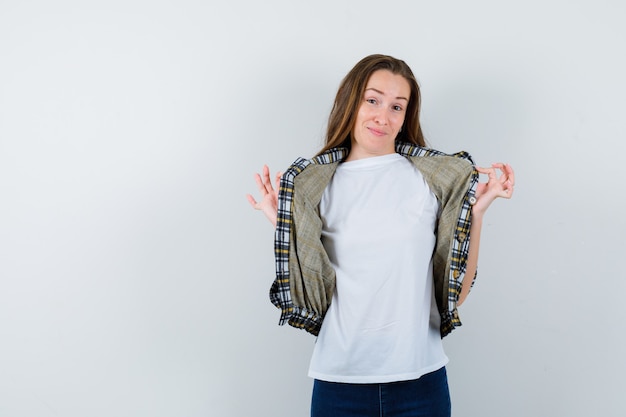 Jovencita en camiseta, chaqueta, jeans sosteniendo la chaqueta con las manos mientras posa y se ve linda, vista frontal.