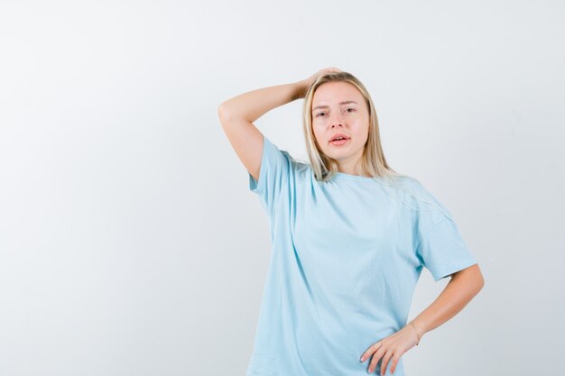 Jovencita en camiseta sosteniendo la mano en la cabeza mientras mantiene la mano en la cintura y mira pensativa, vista frontal.