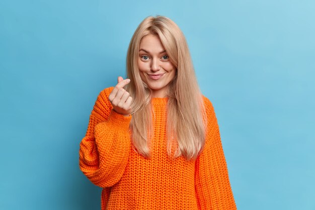 Jovencita caucásica muestra gesto de mano de mini corazón tiene una sonrisa agradable vestida con un jersey naranja casual