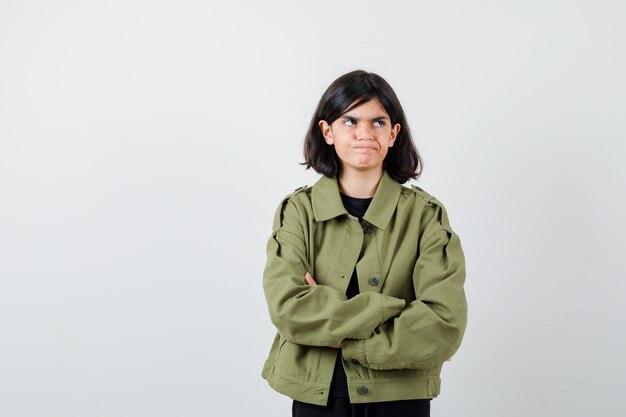 Jovencita en chaqueta verde militar sosteniendo los brazos cruzados, mirando a otro lado y mirando disgustado, vista frontal.