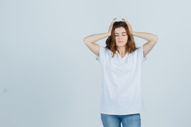 Jovencita cogidos de la mano en la cabeza en camiseta, jeans y con aspecto cansado, vista frontal.