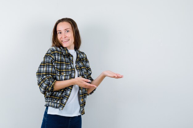 Jovencita dando la bienvenida a algo en camiseta, chaqueta, jeans y luciendo alegre. vista frontal.