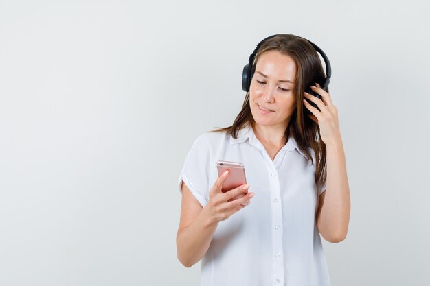 Jovencita escuchando música mientras mira su teléfono en blusa blanca y mira concentrada.