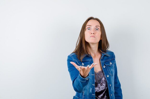 Jovencita estirando la mano en gesto de interrogación en blusa, chaqueta de mezclilla y mirando enojado, vista frontal.