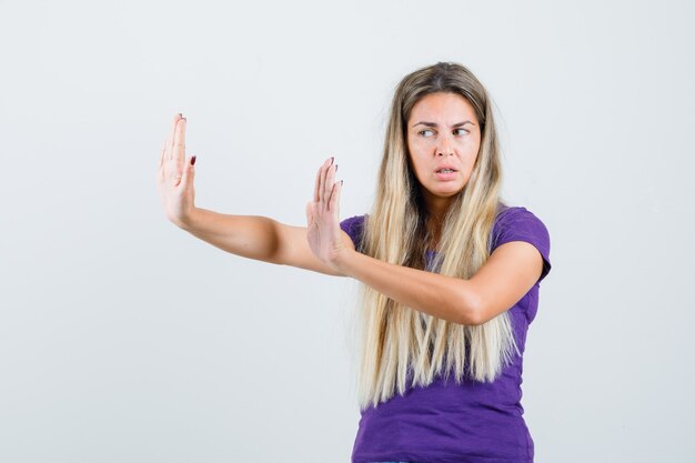 Jovencita estirando las manos para defenderse en camiseta violeta y mirando irritada, vista frontal.