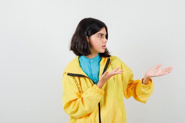 Jovencita fingiendo mostrar algo en chaqueta amarilla y mirando perplejo, vista frontal.