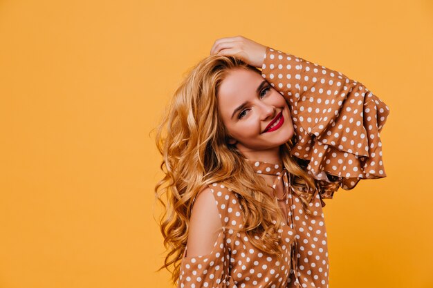 Jovencita juguetona tocando su cabello ondulado y riendo. Filmación en interiores de espectacular modelo femenino en vestido aislado en pared amarilla.