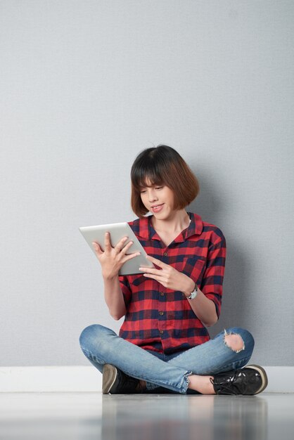 Jovencita leyendo un libro electrónico interesante sentado en postura de loto en el piso de su habitación del campus